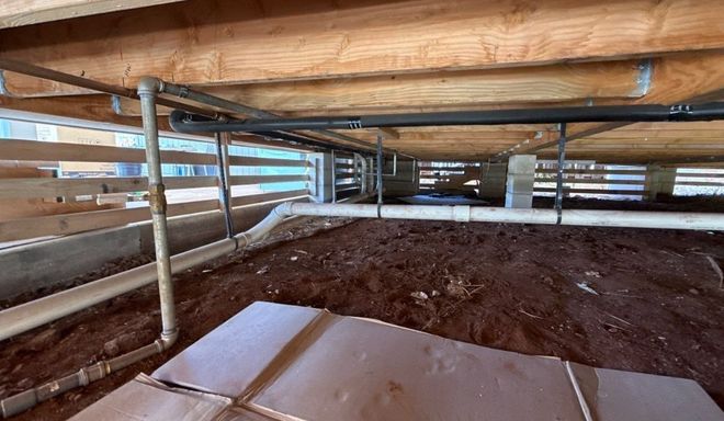 Bare crawl space with exposed beams, pipes, and dirt floor under a house.