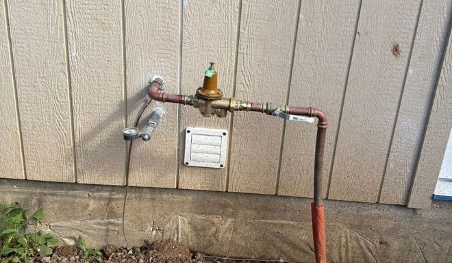 Outdoor water meter and shutoff valve on a house wall with pipes and a hose attached.