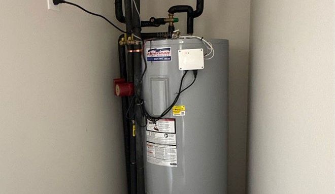 Gray water heater with pipes and control valves in a utility closet against a white wall.