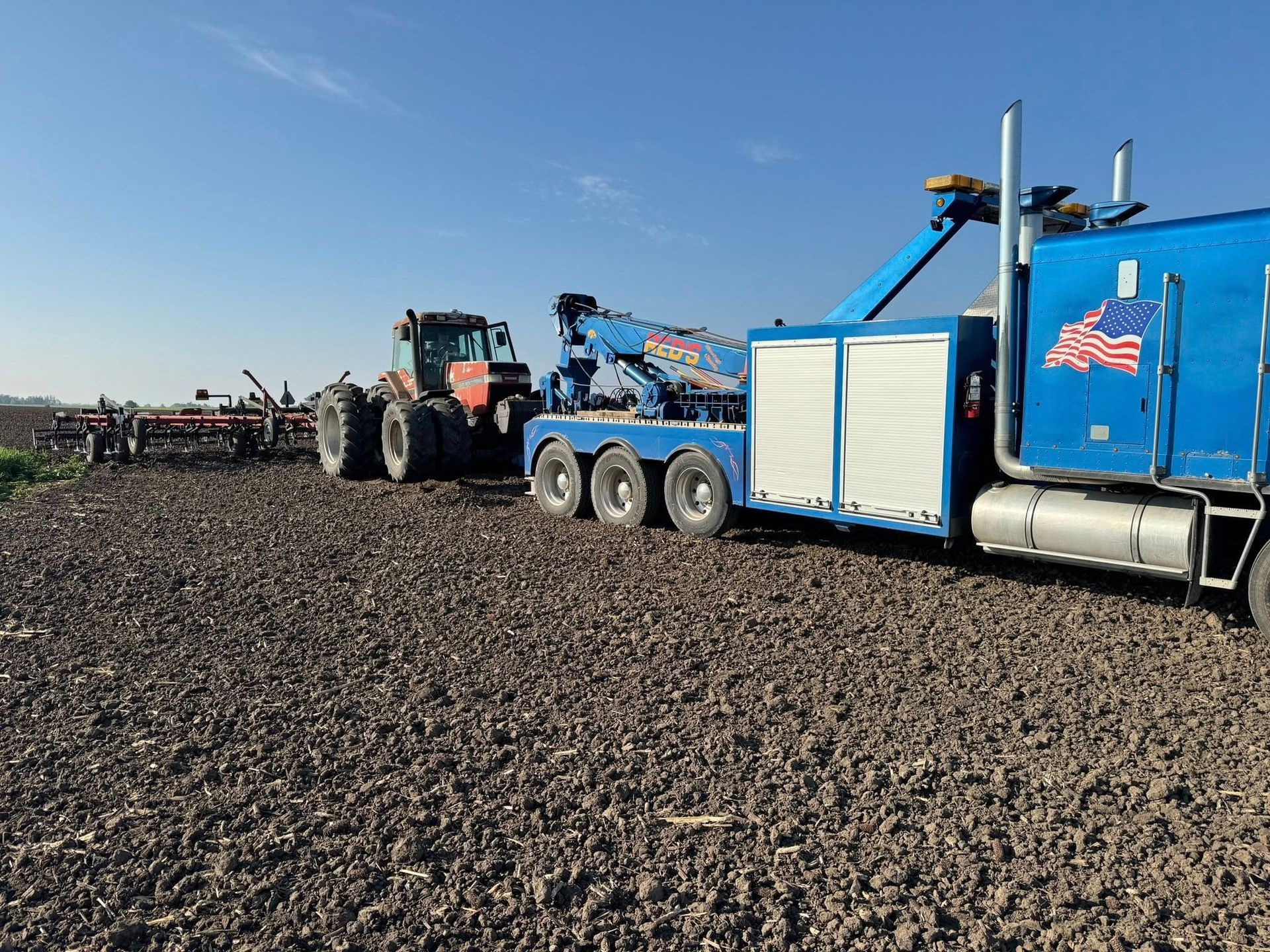 A blue tow truck pulling a tractor in a plowed field under a clear sky.