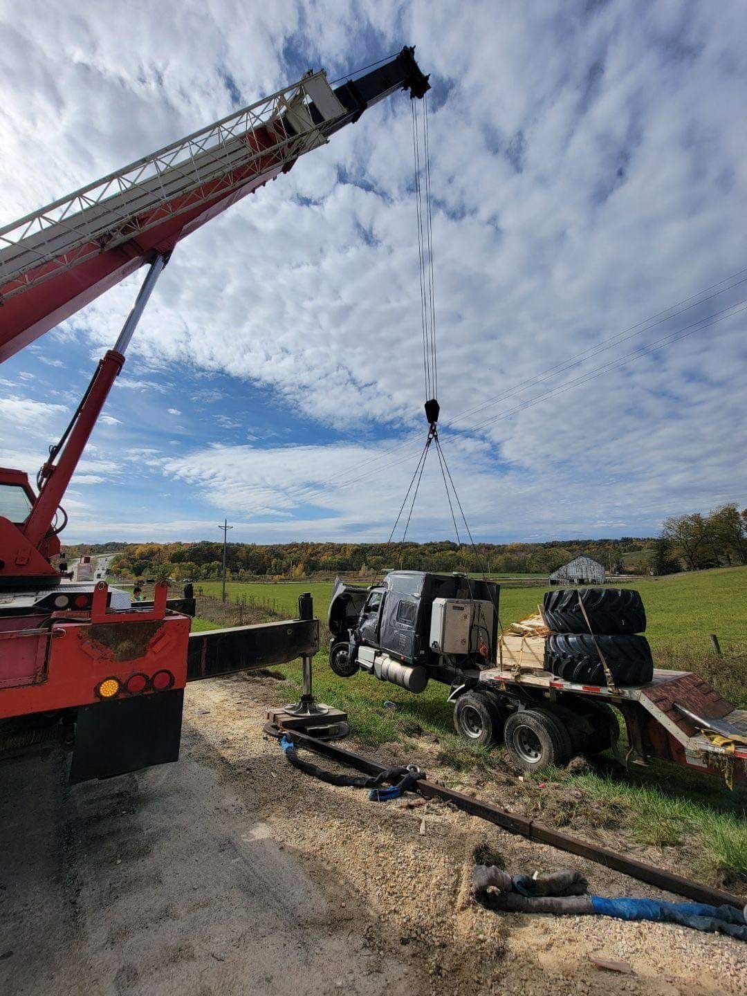 Crane lifting a truck from a trailer on a gravel road, fields in the background. Cloudy sky.
