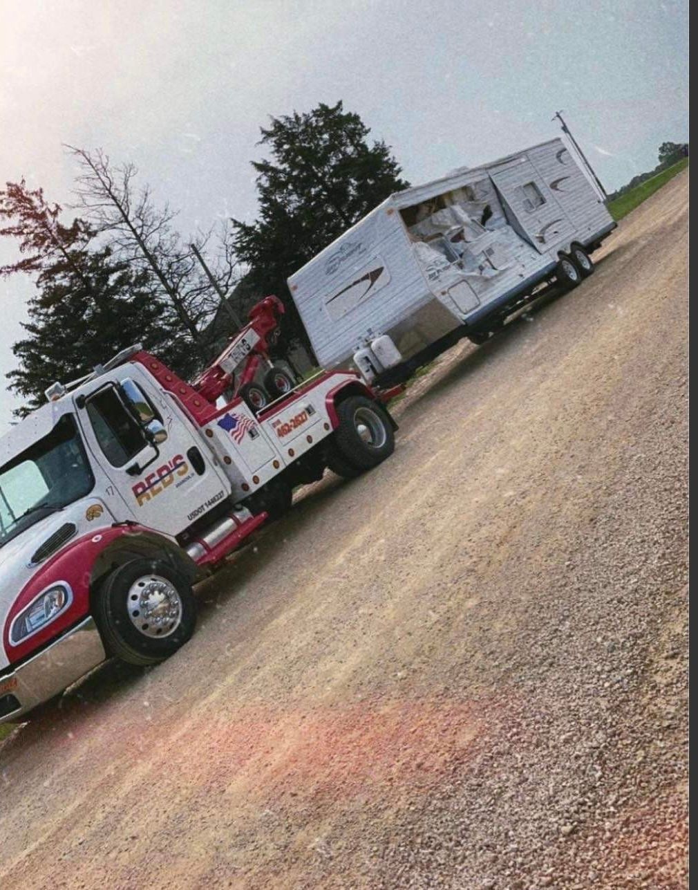 Tow truck towing a travel trailer on a gravel road, overcast sky.