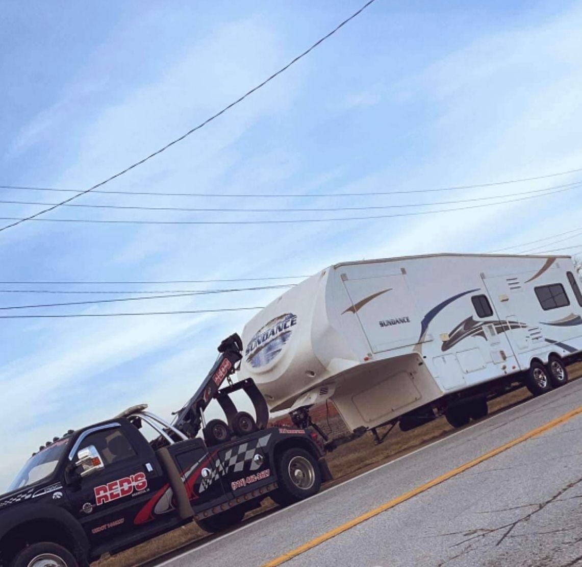 A tow truck is towing a white RV on a road under a blue sky.