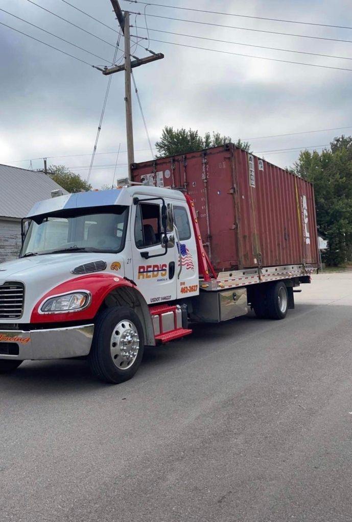 A red shipping container on a flatbed truck driving down a street. The truck is white, red, and blue.
