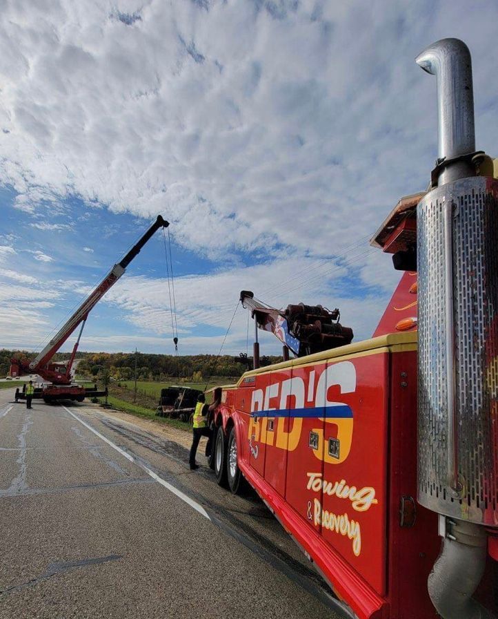 Tow truck next to highway, crane lifting vehicle, two people in vests assisting under cloudy sky.