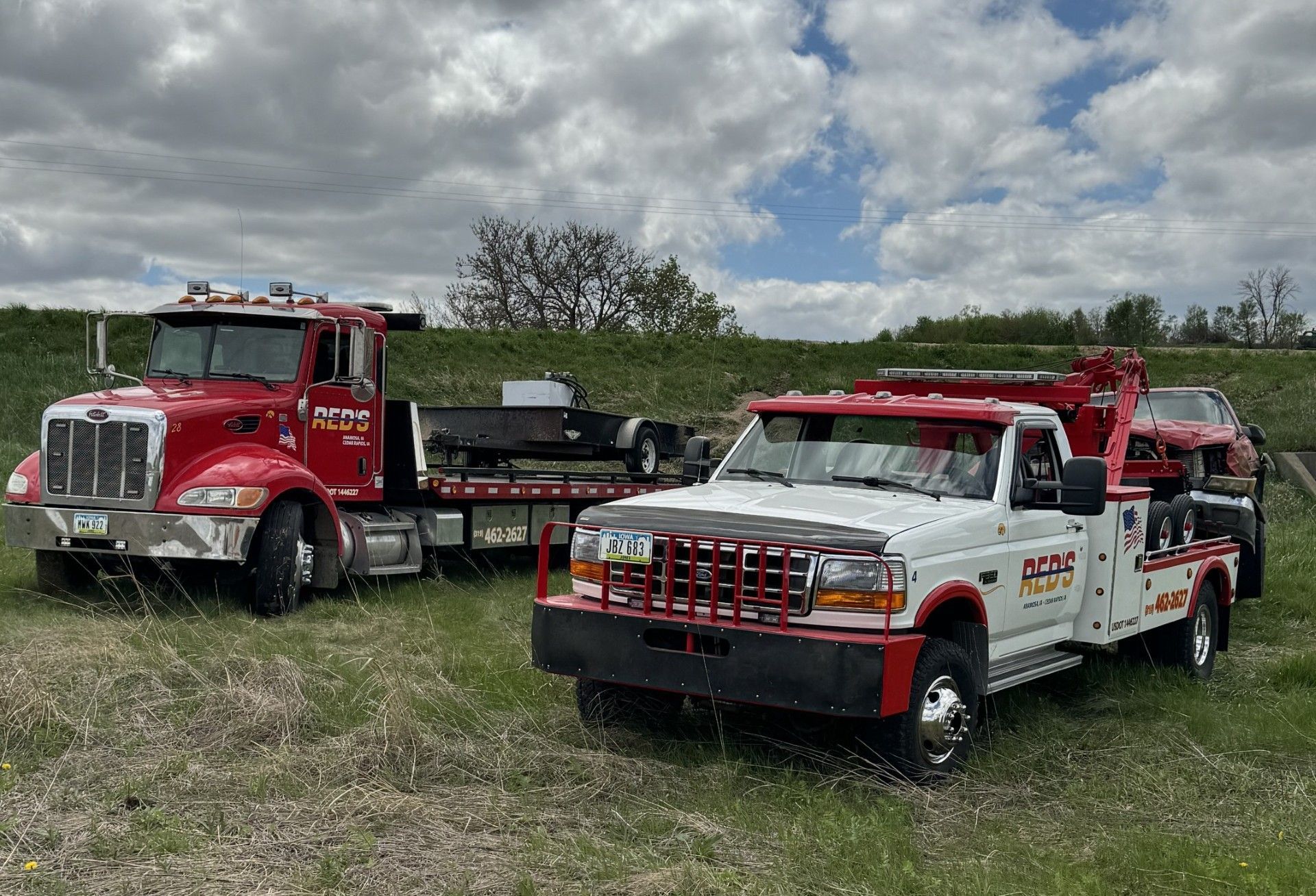 Two tow trucks, red and white, parked on grassy land under a cloudy sky.