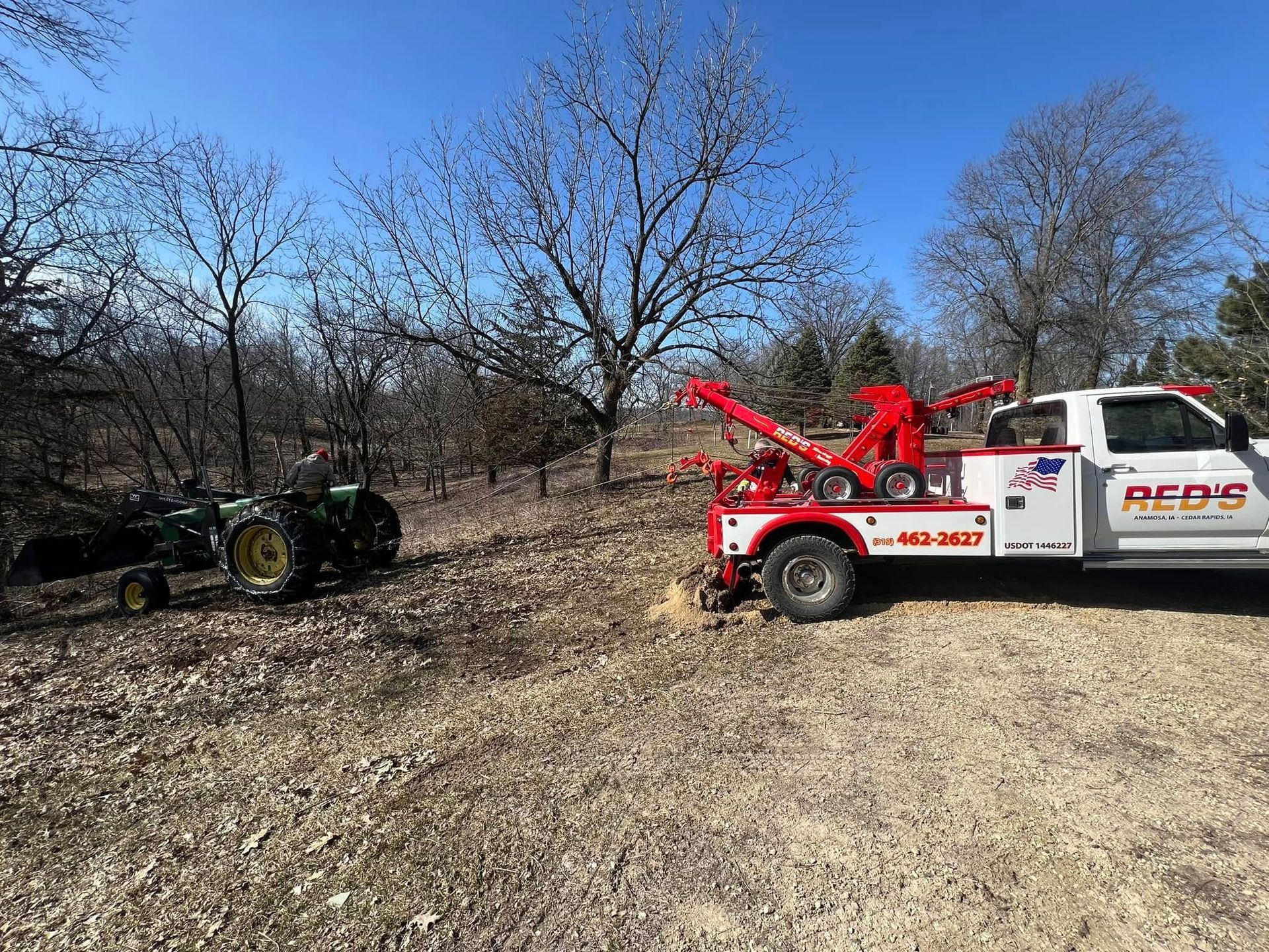A tow truck is pulling a green tractor from a wooded area on a sunny day.