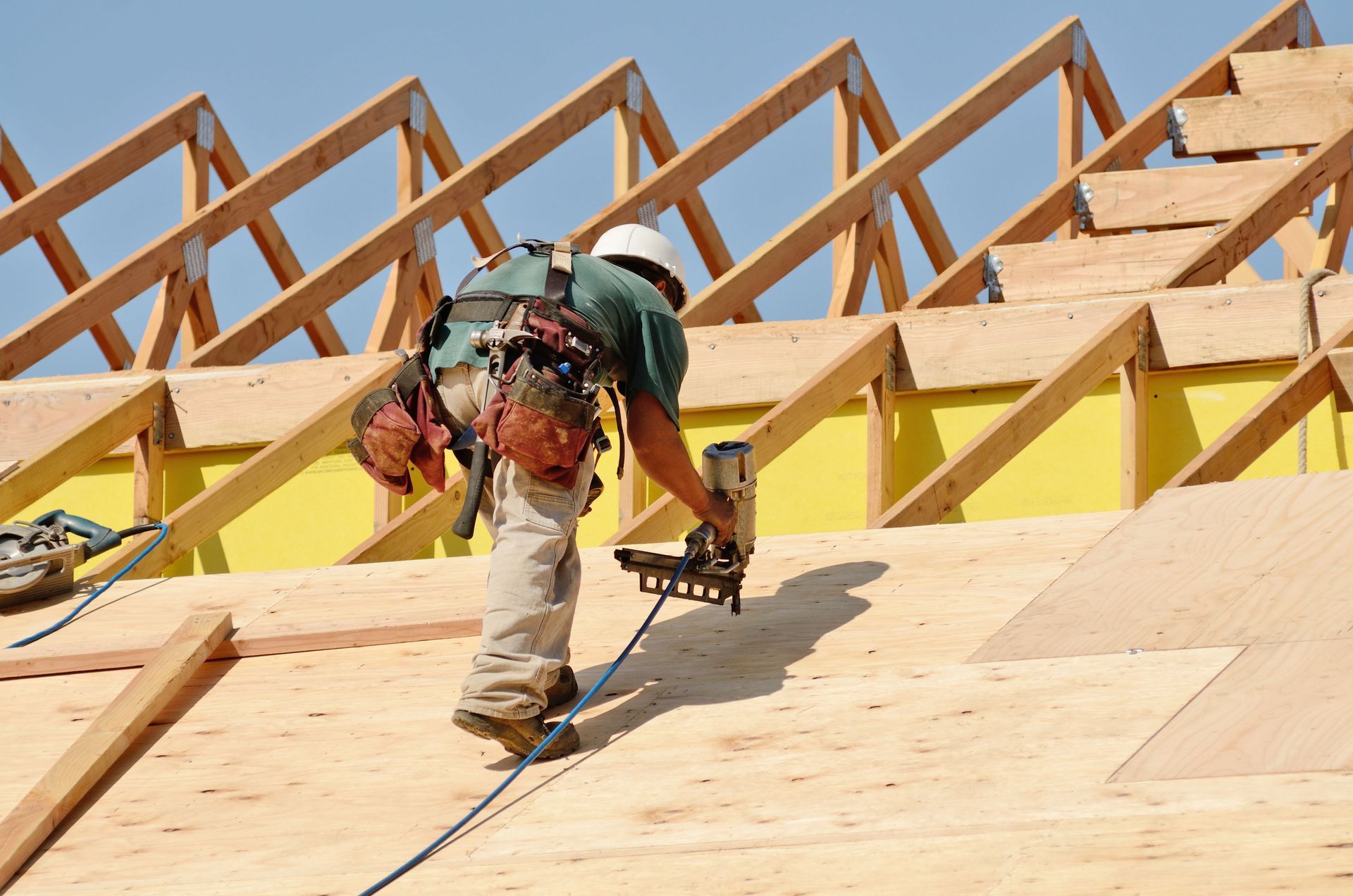 Construction worker on a roof nailing plywood. Wearing safety harness, wood frame visible, blue sky.