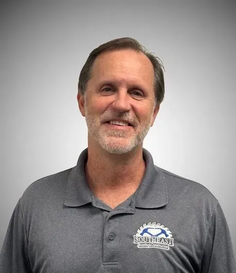 Man with salt and pepper hair and beard, wearing a gray polo shirt with a logo, smiling, indoors.