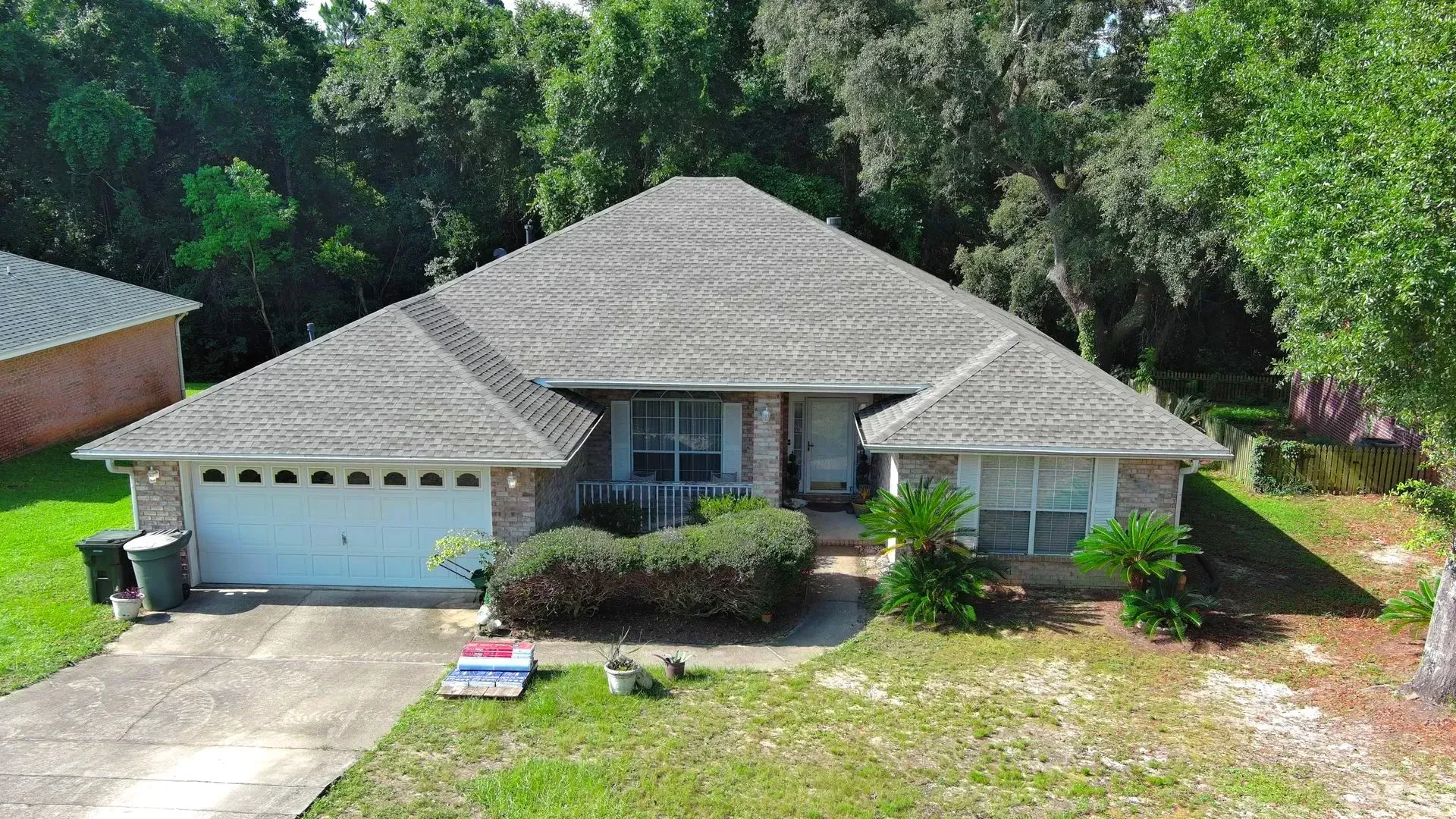 Residential house with gray roof, brick facade, and attached garage. Green lawn, trees in background.