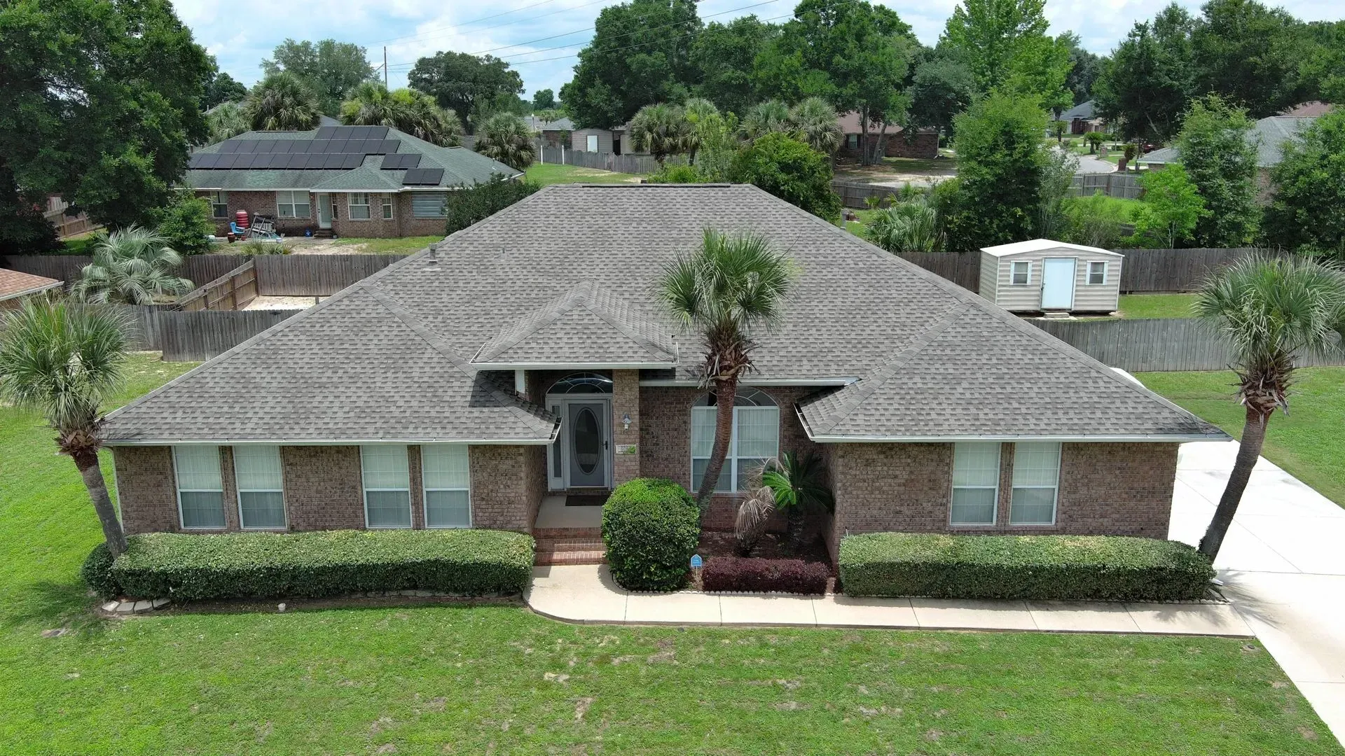 Brick house with gray roof, green lawn, palm trees, and bushes in front.