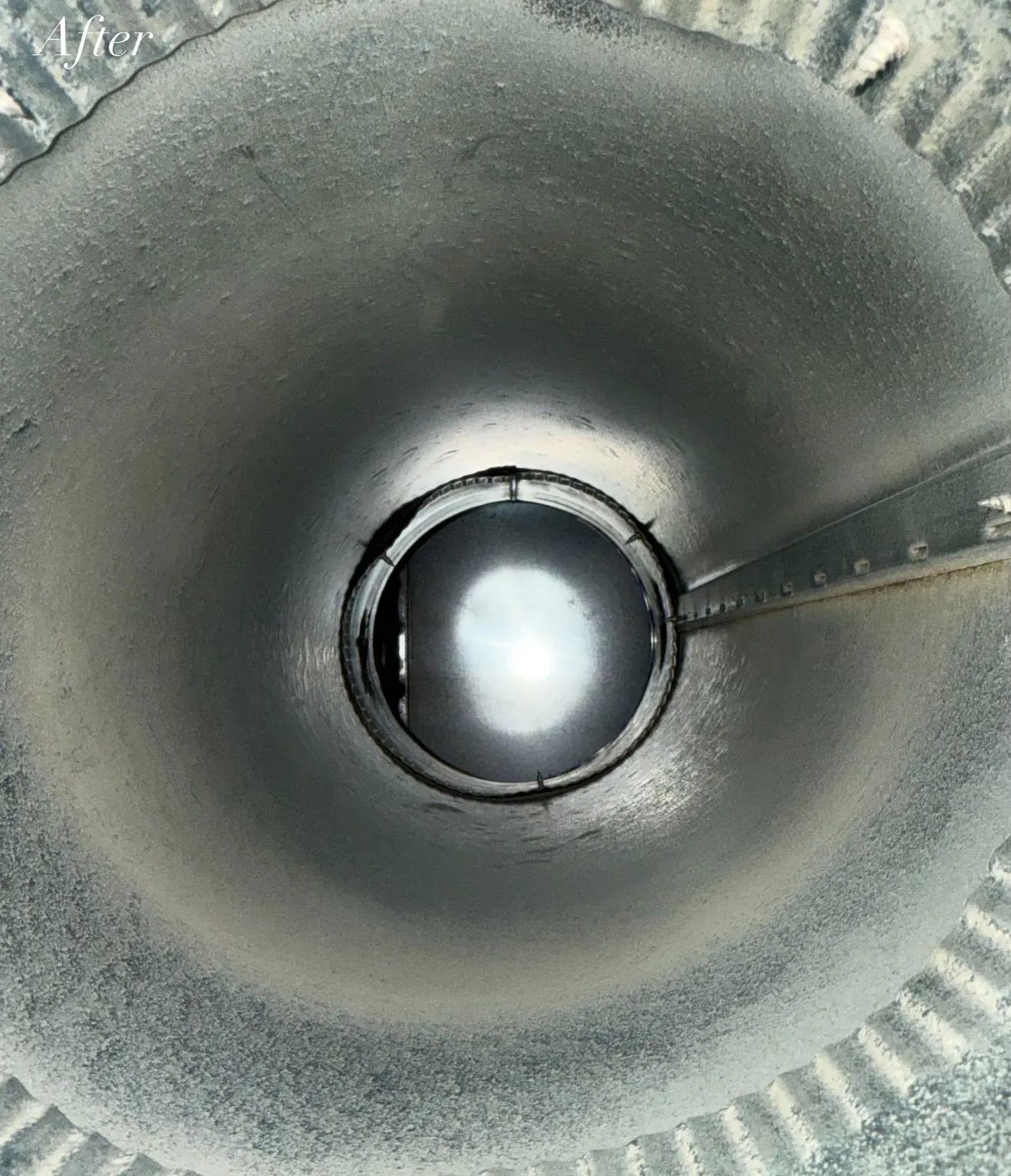 Inside view of a metallic, dusty air duct. Light at the center, visible seam and ribs on outer edge.