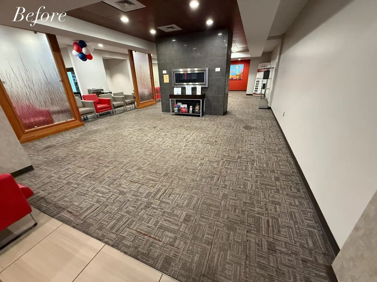 Waiting area with gray patterned carpet, red chairs, and a TV stand. Brown and white walls.