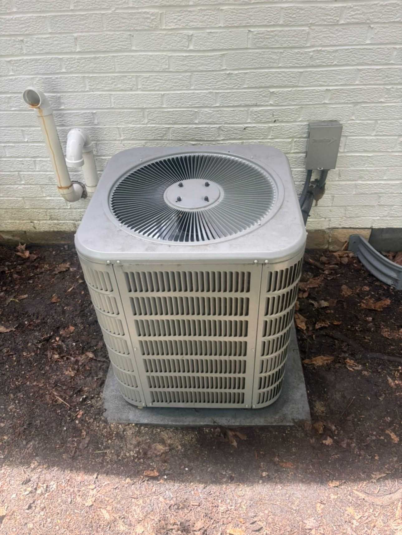 Outdoor air conditioning unit against a white brick wall, with a fan and electrical box.