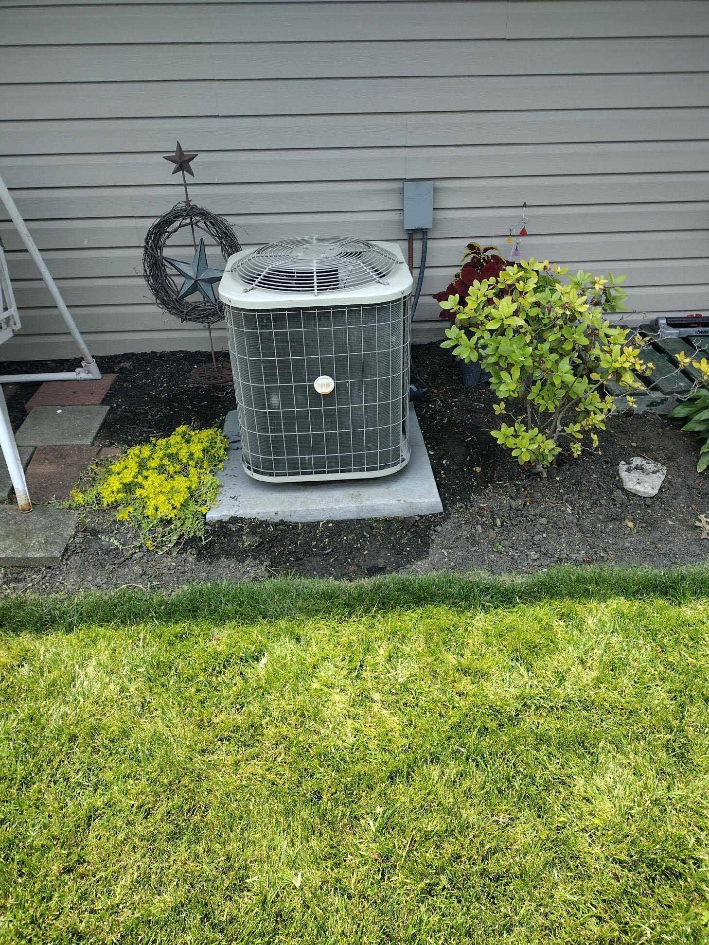 Outdoor air conditioning unit with damaged top, set on a concrete pad, surrounded by a garden bed and grass.