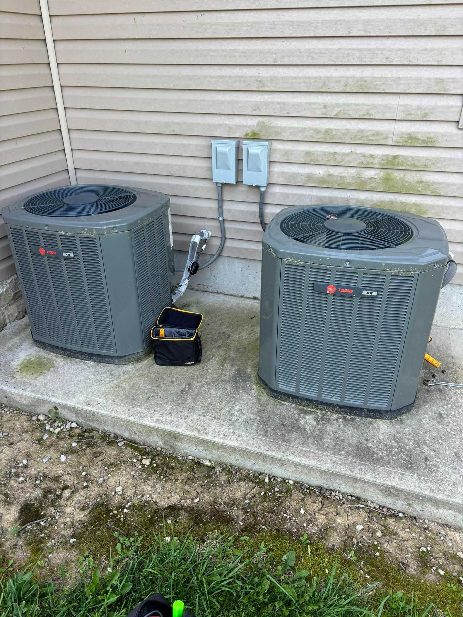 Two gray air conditioning units on a concrete pad near a wall with electrical boxes.