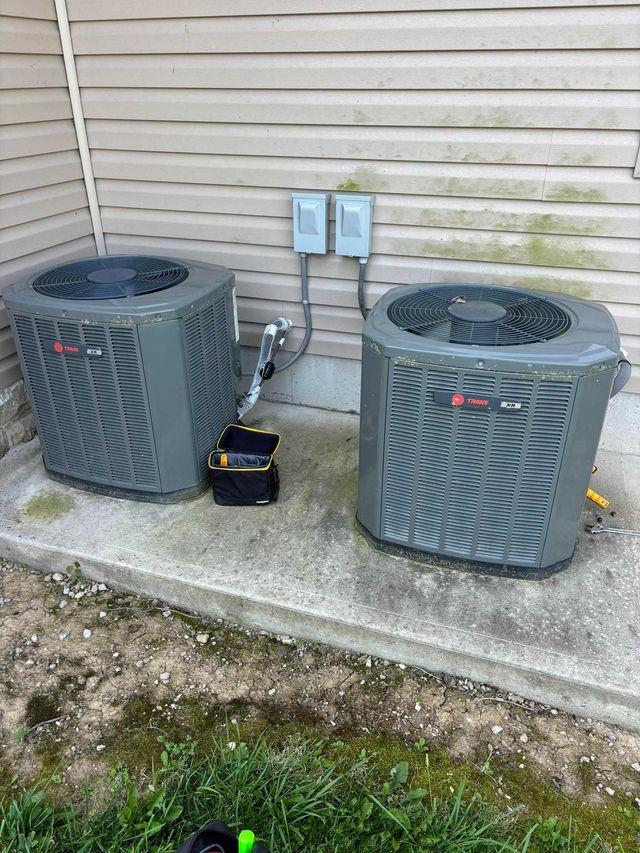 Two gray air conditioning units on a concrete pad near a wall with electrical boxes.