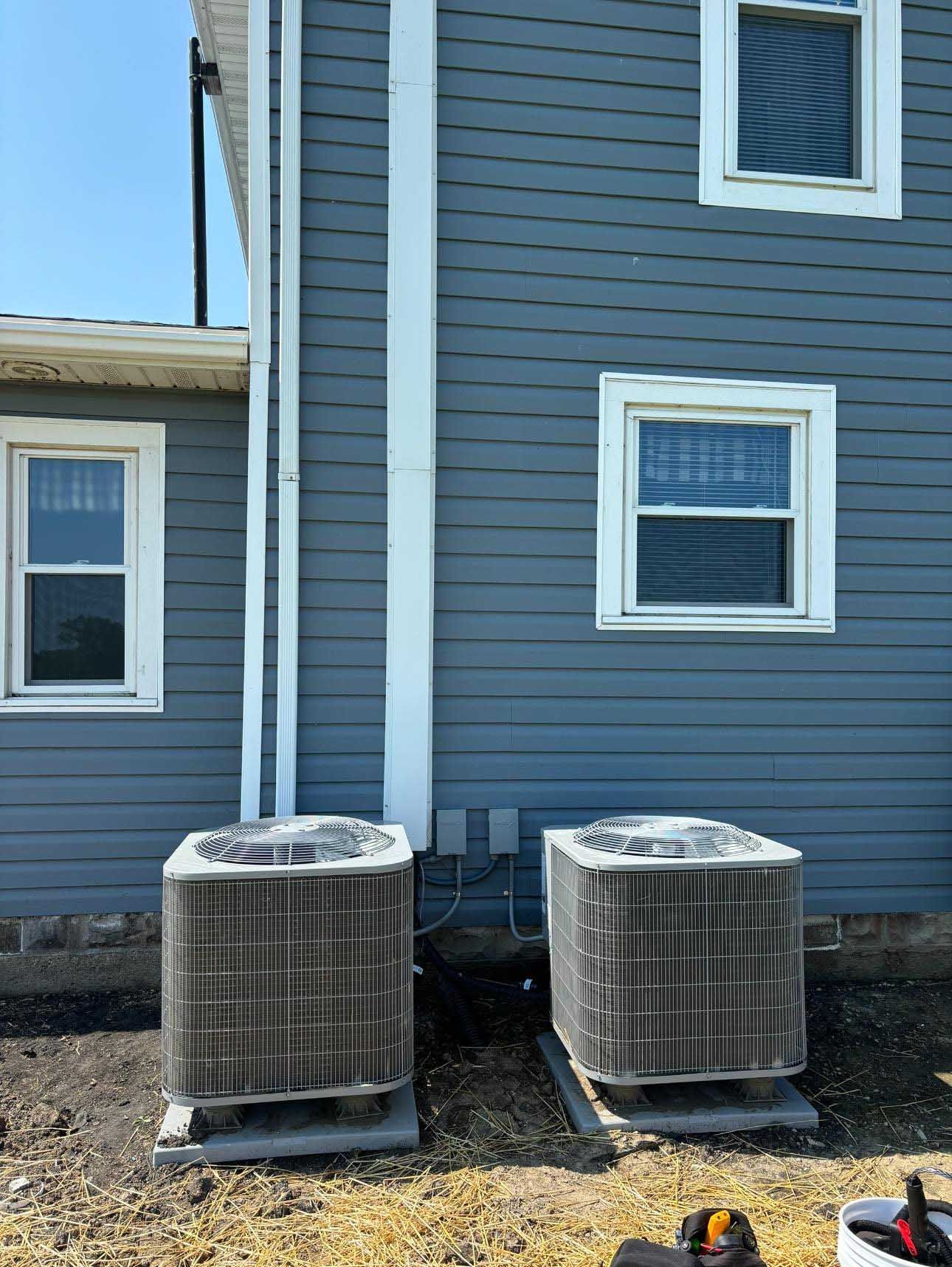 Two air conditioning units sit next to a blue house with white trim windows and a downspout.