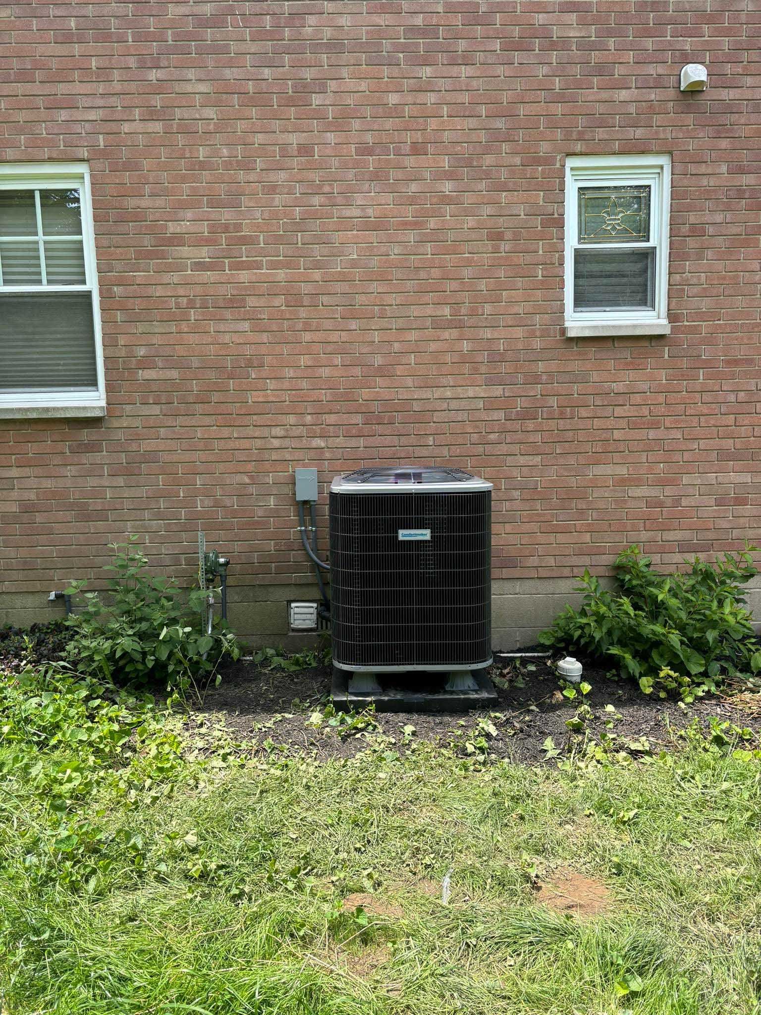 Air conditioning unit against a brick wall, flanked by bushes and windows.