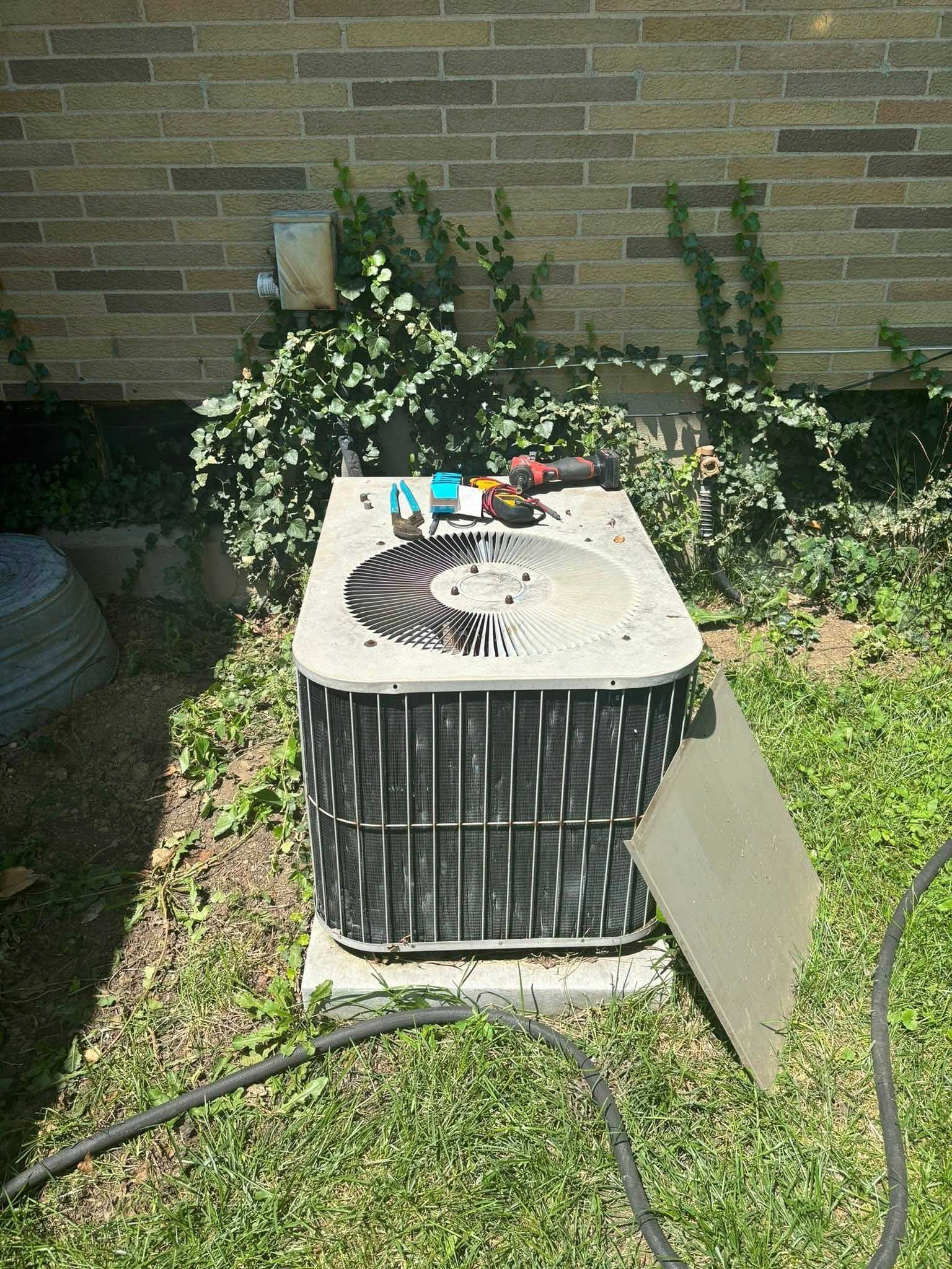 An outdoor air conditioning unit with tools on top, sitting in grass next to a brick wall with vines.