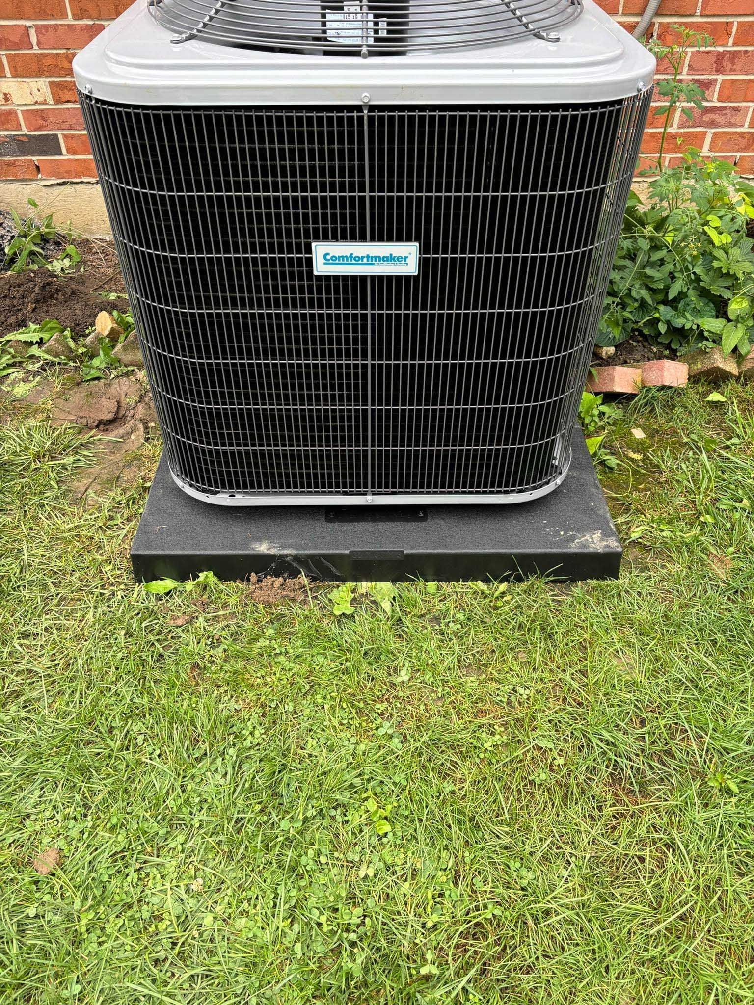 An air conditioning unit on a dark pad in a grassy yard, near a brick wall.
