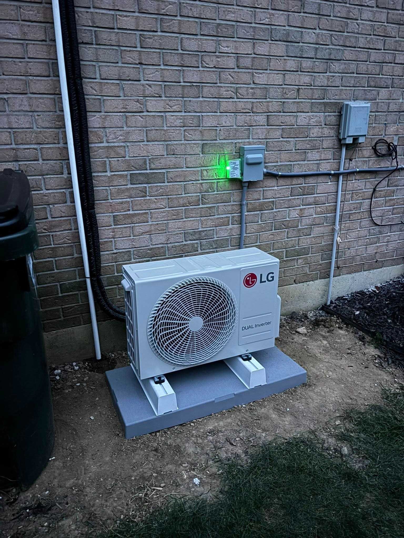 LG heat pump unit on a concrete pad near a brick wall with electrical components. Green light is on.