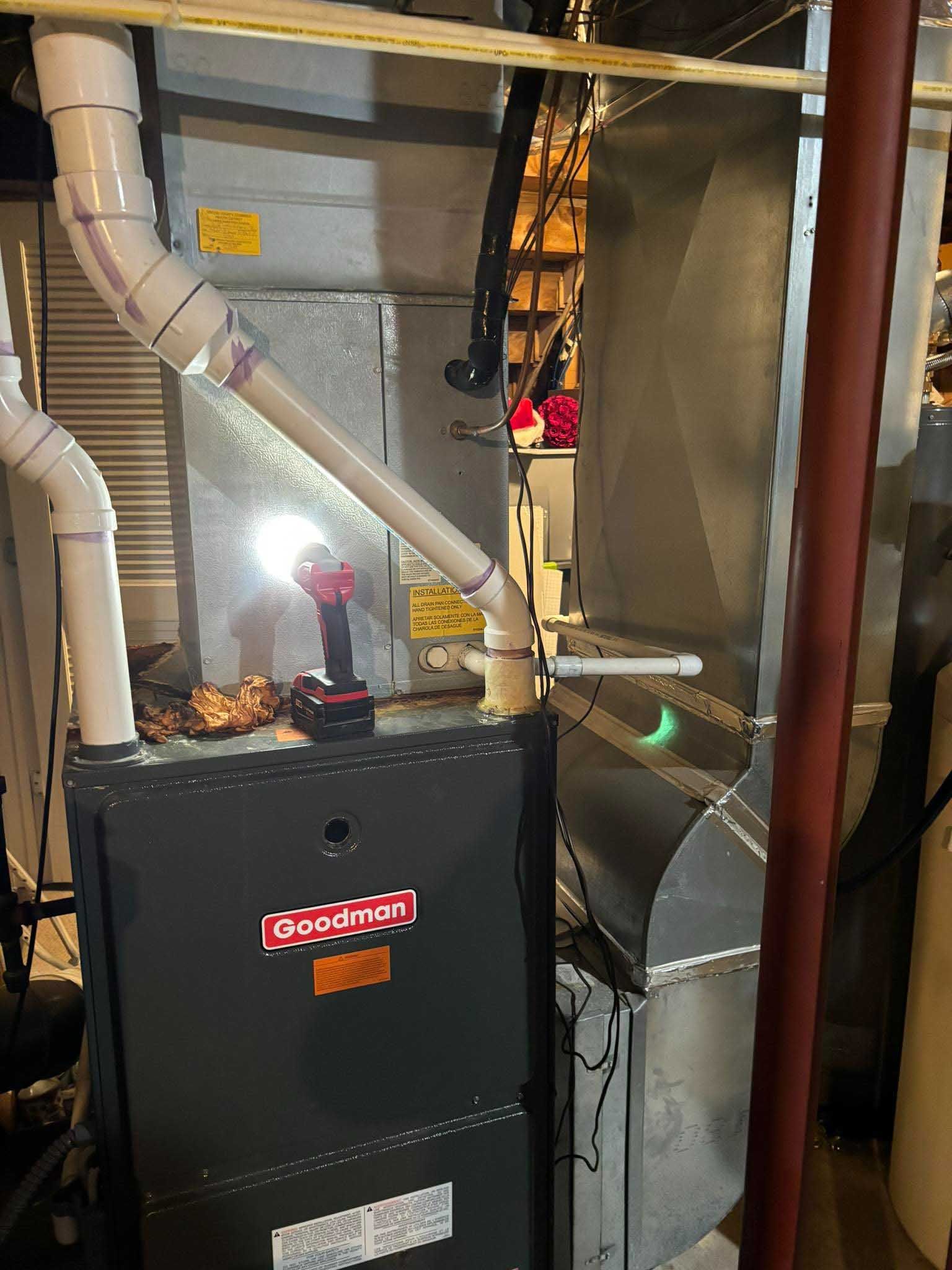 A Goodman furnace with attached ductwork and PVC pipes; a power drill rests on top.