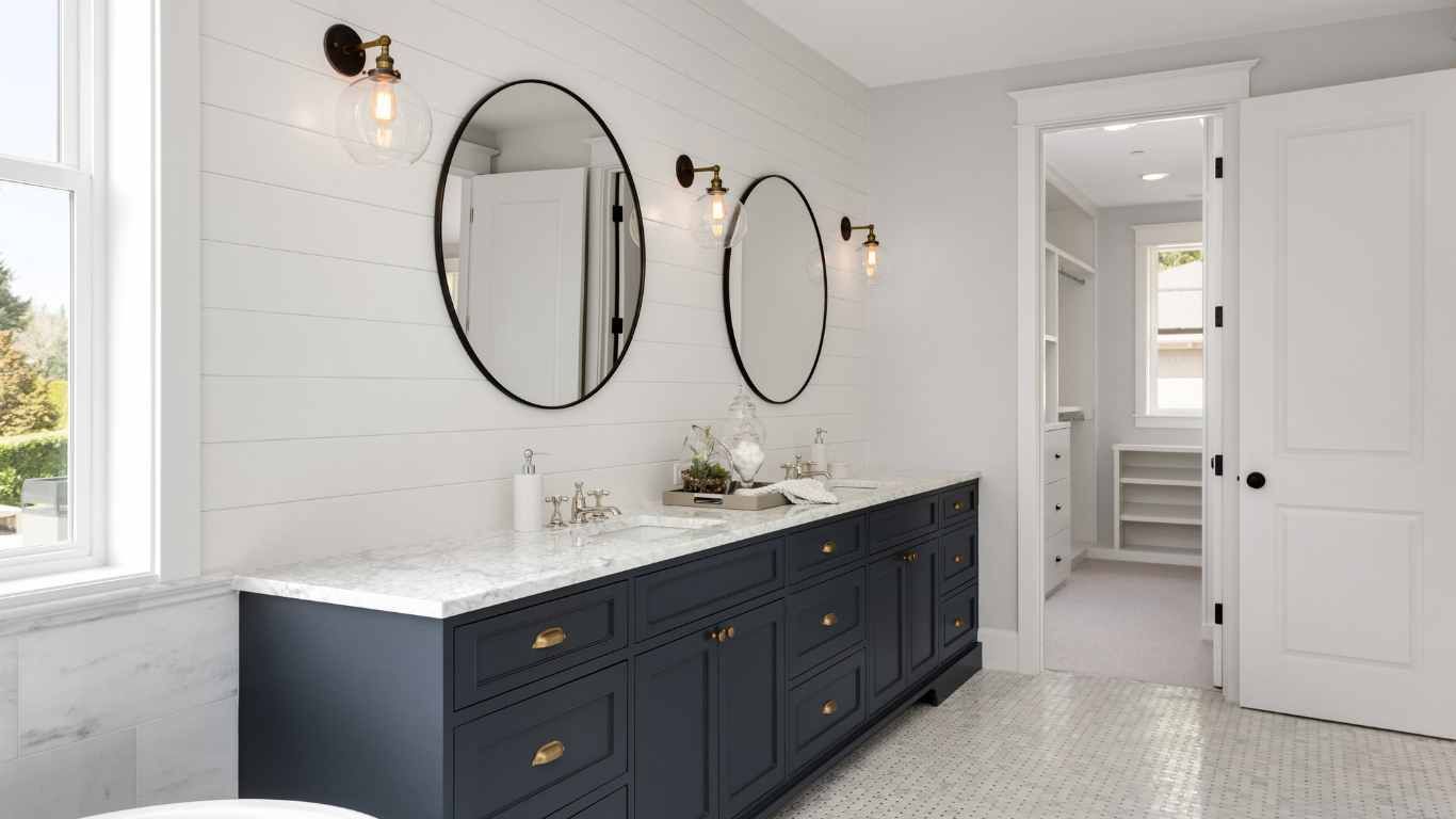 Bathroom with a dark blue vanity, two oval mirrors, and white shiplap walls. A door leads to a closet.