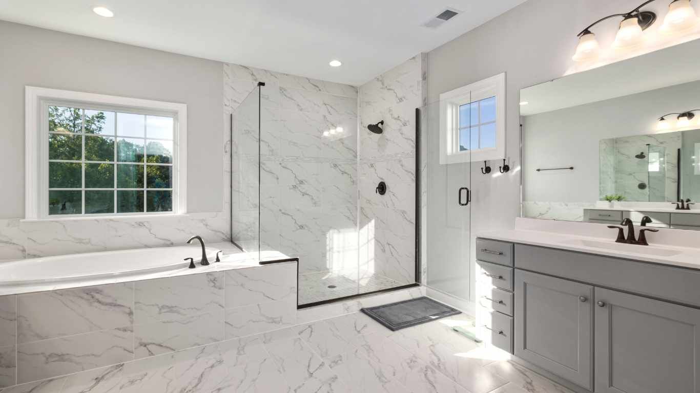 Spacious modern bathroom with marble tile, glass shower, gray vanity, and soaking tub by window.