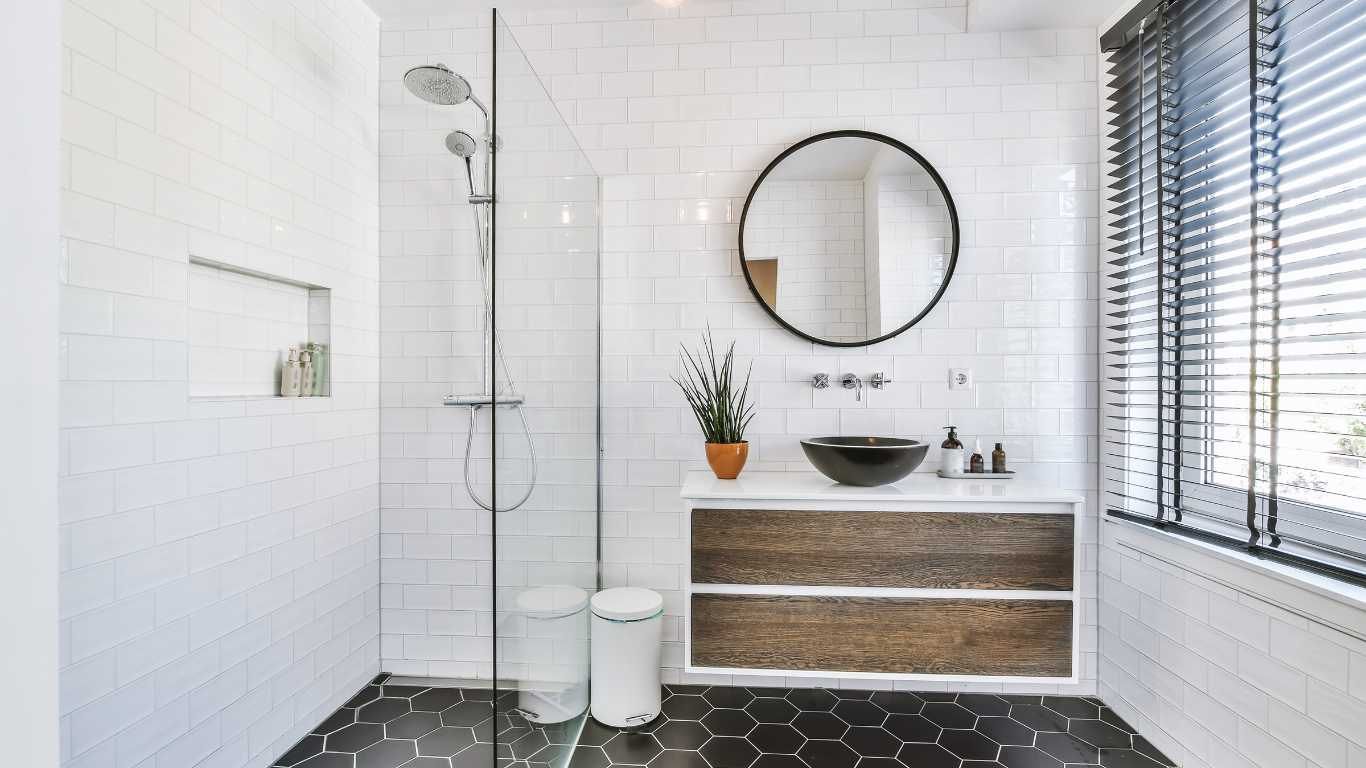 Modern bathroom with black hexagon tiles, white subway tile walls, glass shower, wooden vanity, and round mirror.