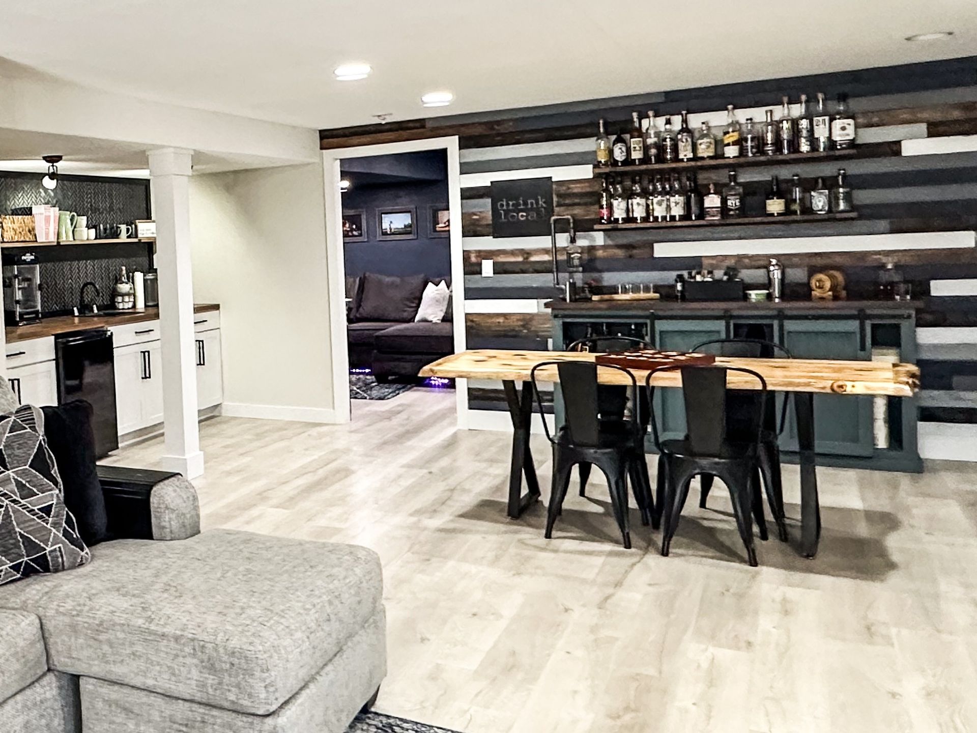 Basement bar area with wooden accent wall, shelves of bottles, table with chairs, and sectional sofa.