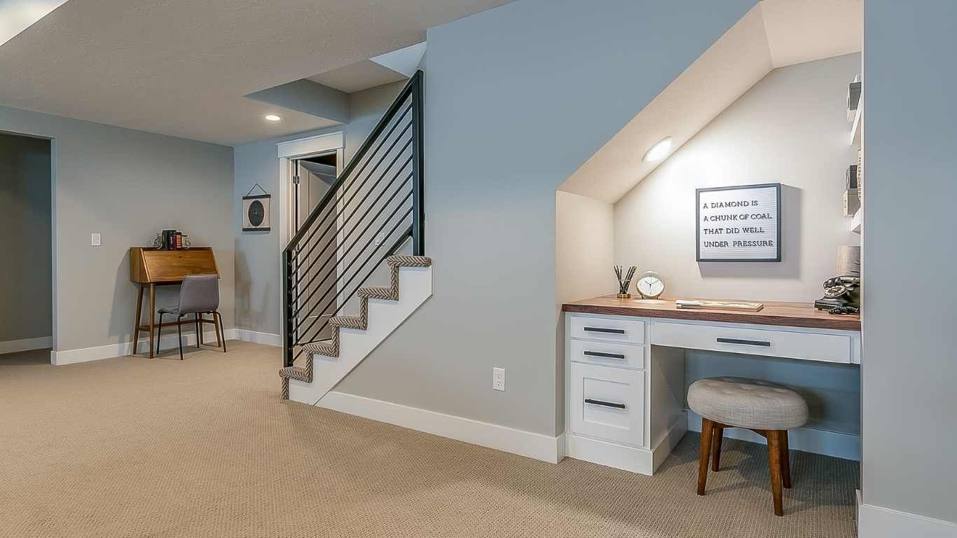 Basement with built-in desk under stairs, stairs with metal railing, beige carpet, small desk in the background.