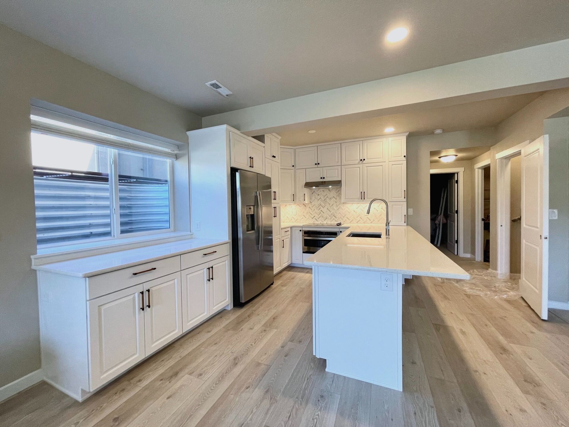 Bright white kitchen with a window, cabinets, island, and stainless steel appliances; light wood flooring.