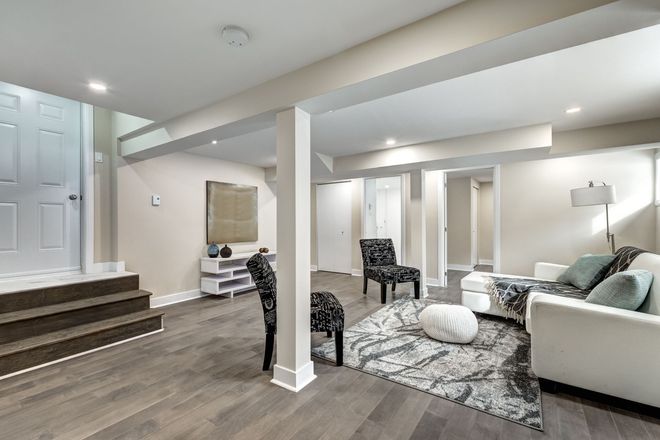 A finished, neutrally-toned basement featuring light wood floors, a sofa, area rug, accent chairs, and wooden stairs.