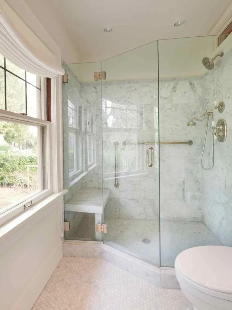 Glass-enclosed shower in a marble-tiled bathroom with a window and a toilet.