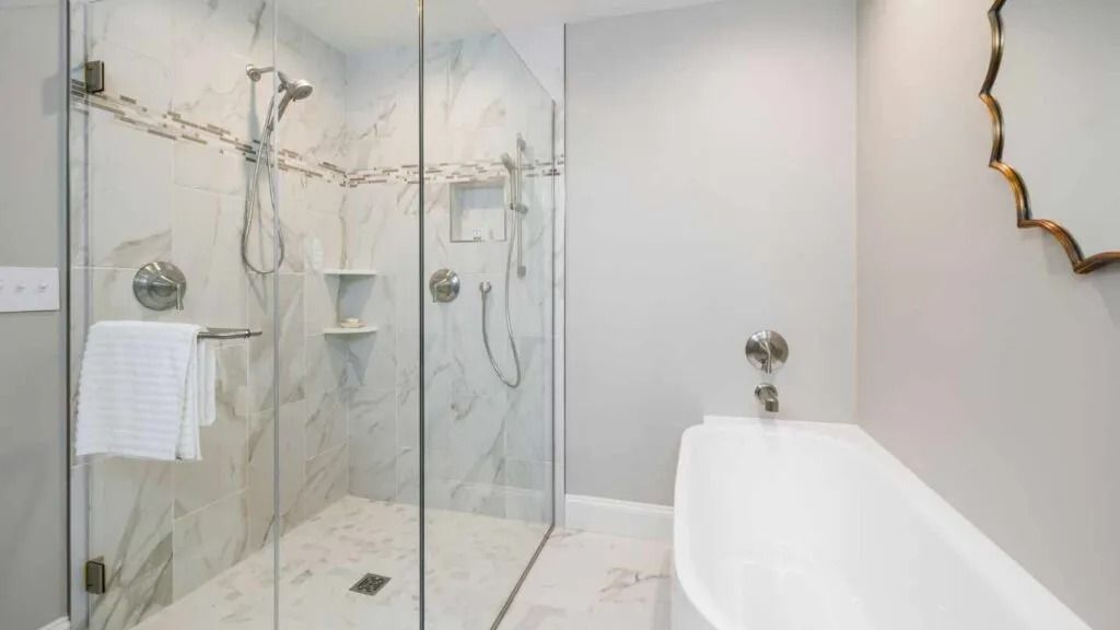 Bathroom with a glass shower, white bathtub, and gold-framed mirror. Shower has marble-like tile and chrome fixtures.