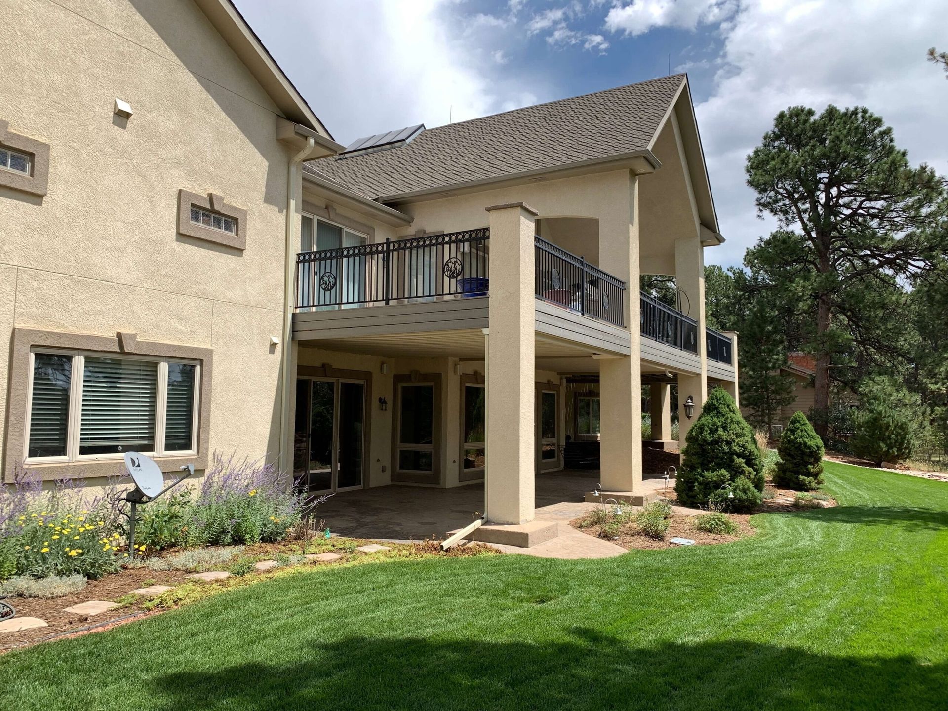 Back of a two-story beige house with a covered patio, balcony with black railings, and a green lawn.