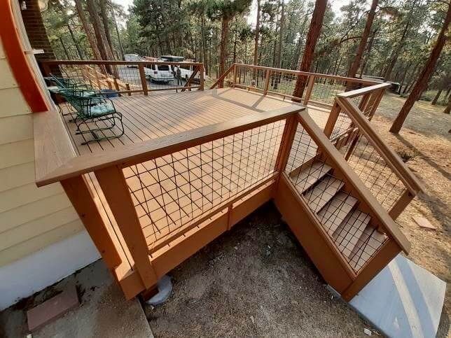 Wooden deck with stairs, surrounded by trees. Tan railing with metal wire mesh.