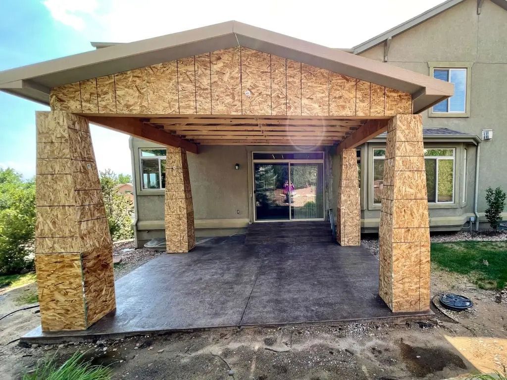 Patio cover under construction: OSB-sheathed supports and roof, concrete patio, home in background.