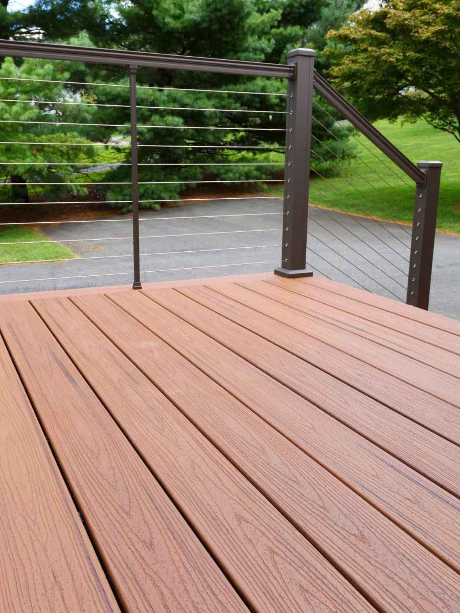 Brown composite deck with cable railing overlooking a paved driveway and green lawn.