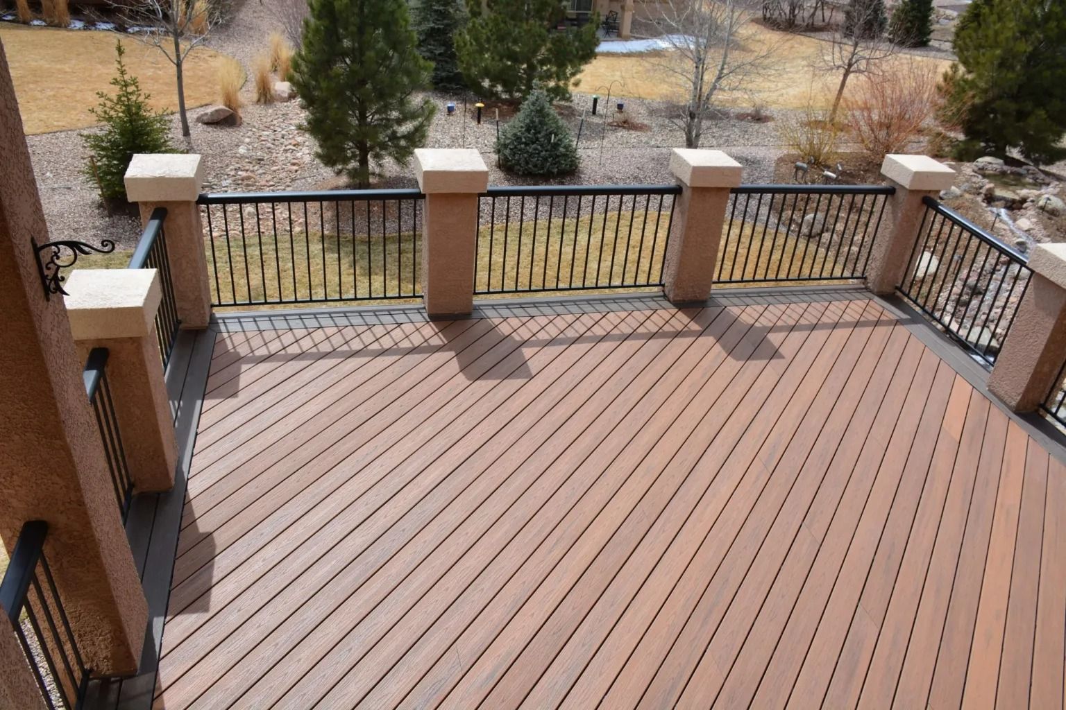 Wooden deck with black railings and light brown pillars overlooking a yard with trees.