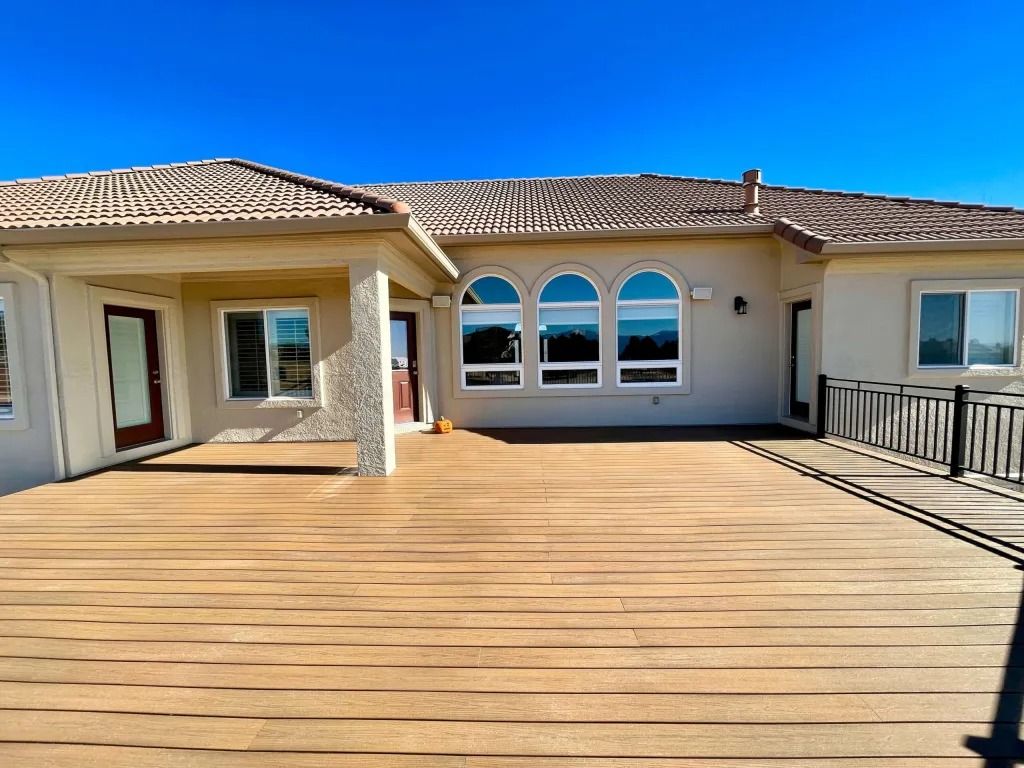 Back patio of a beige house with a brown deck, arched windows, and a tiled roof against a blue sky.