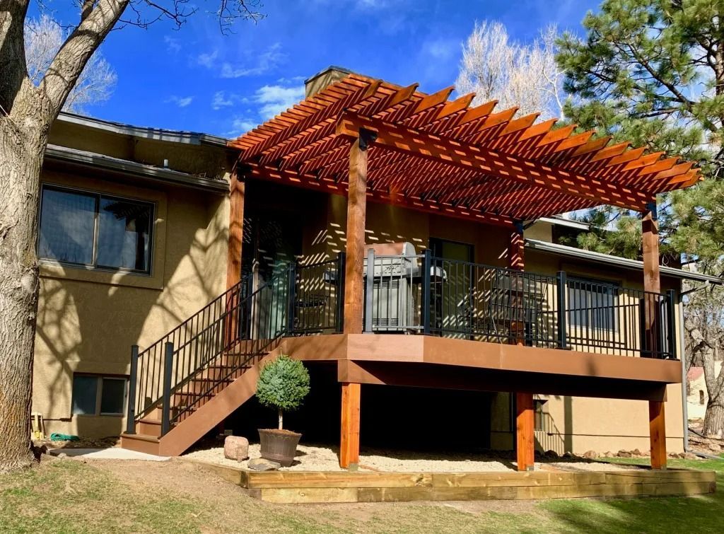Brown wooden deck with pergola on a beige house, with black railings, against a blue sky.