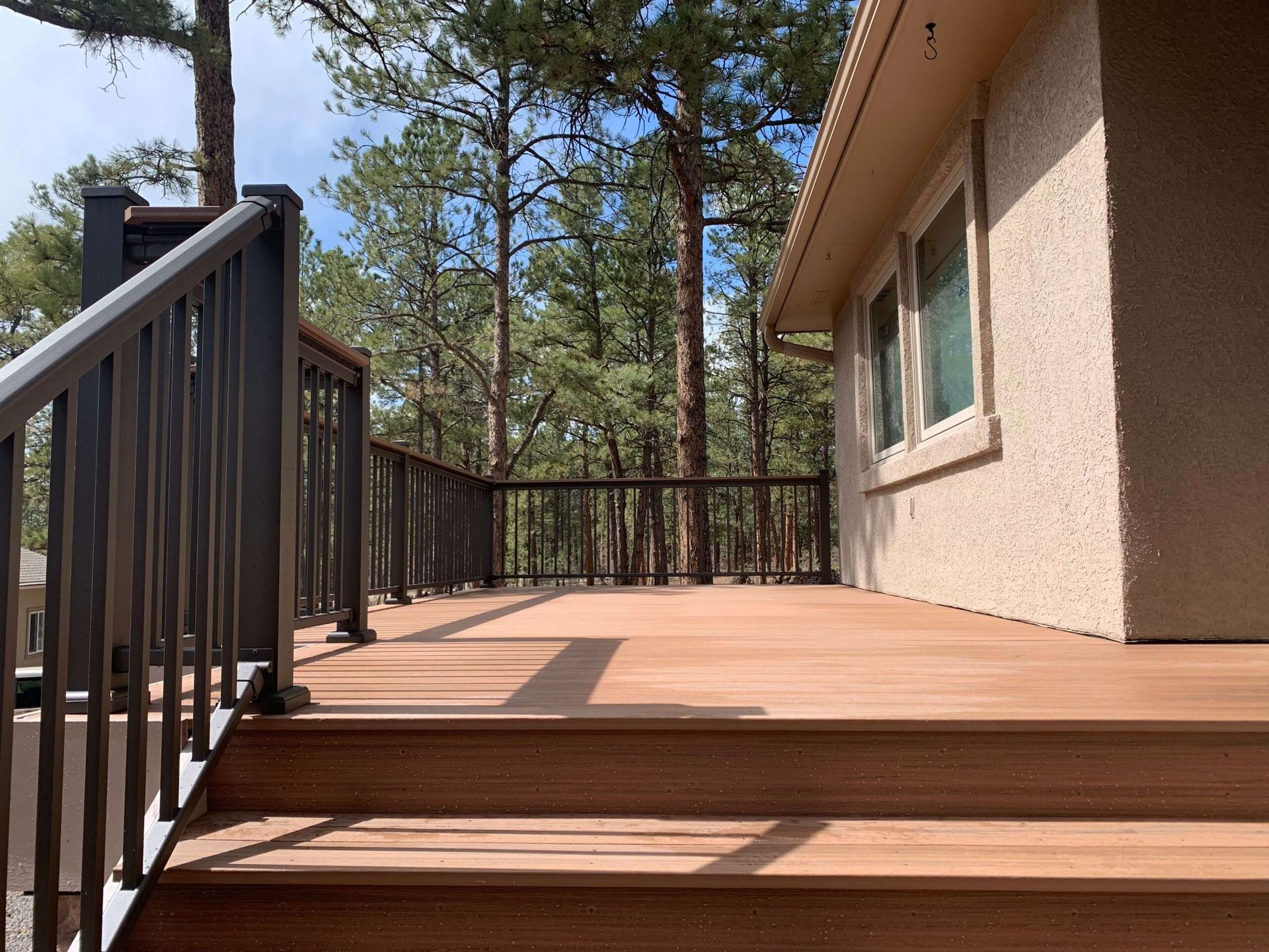Wooden deck with dark railing, adjacent to a beige house, surrounded by trees.