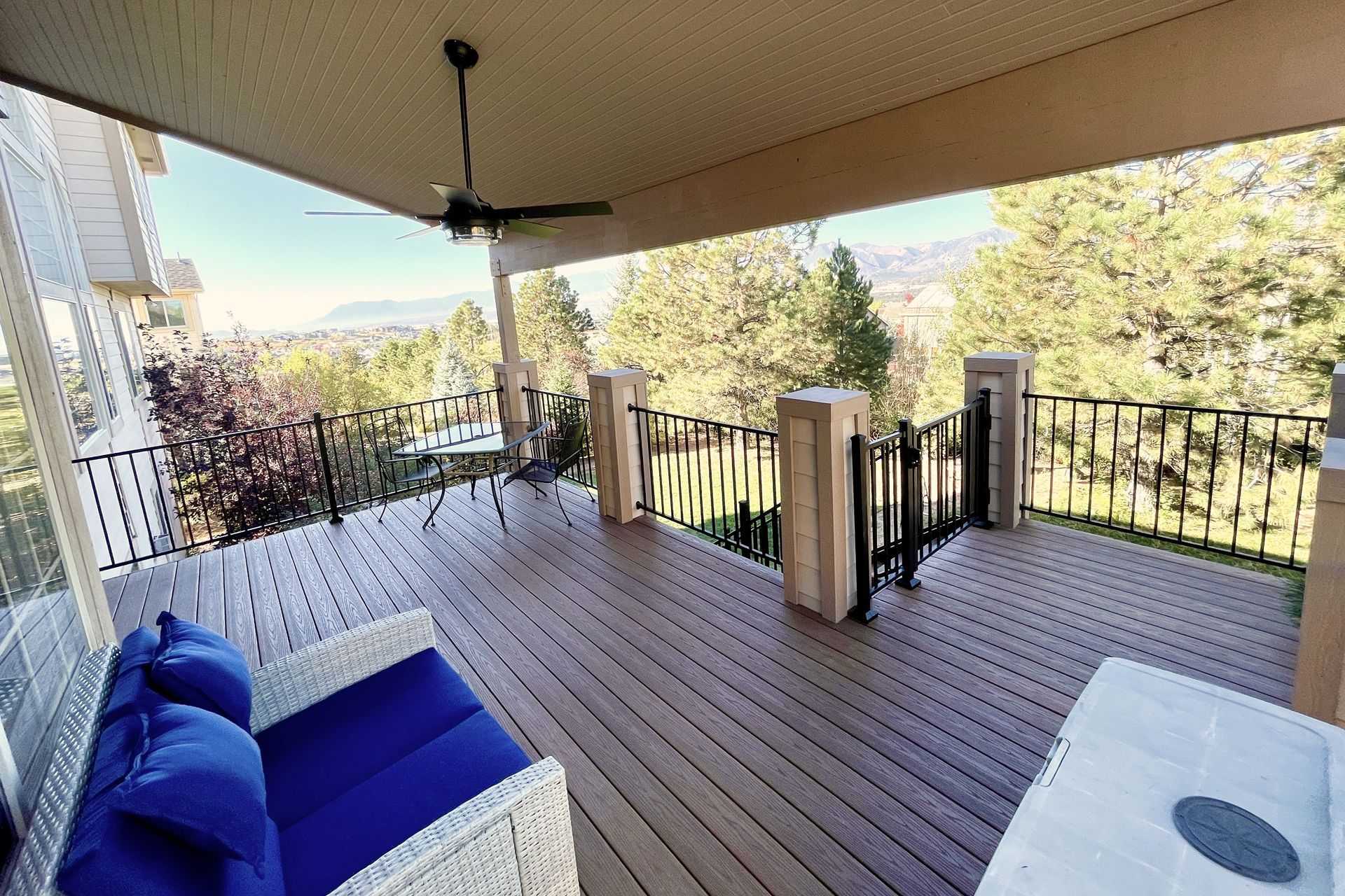 A wooden deck with a covered ceiling fan, patio furniture, and a metal railing overlooking trees and distant mountains.