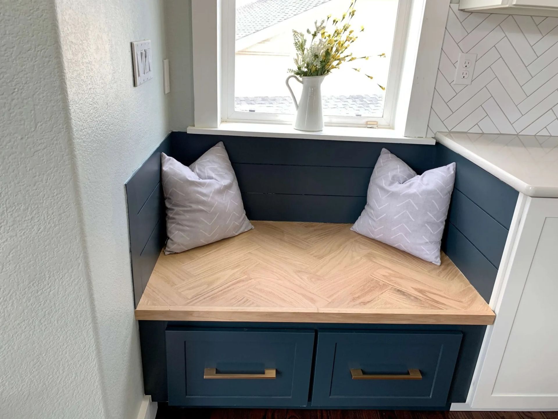 Built-in blue storage bench with herringbone seat, two pillows, and window with flowers.