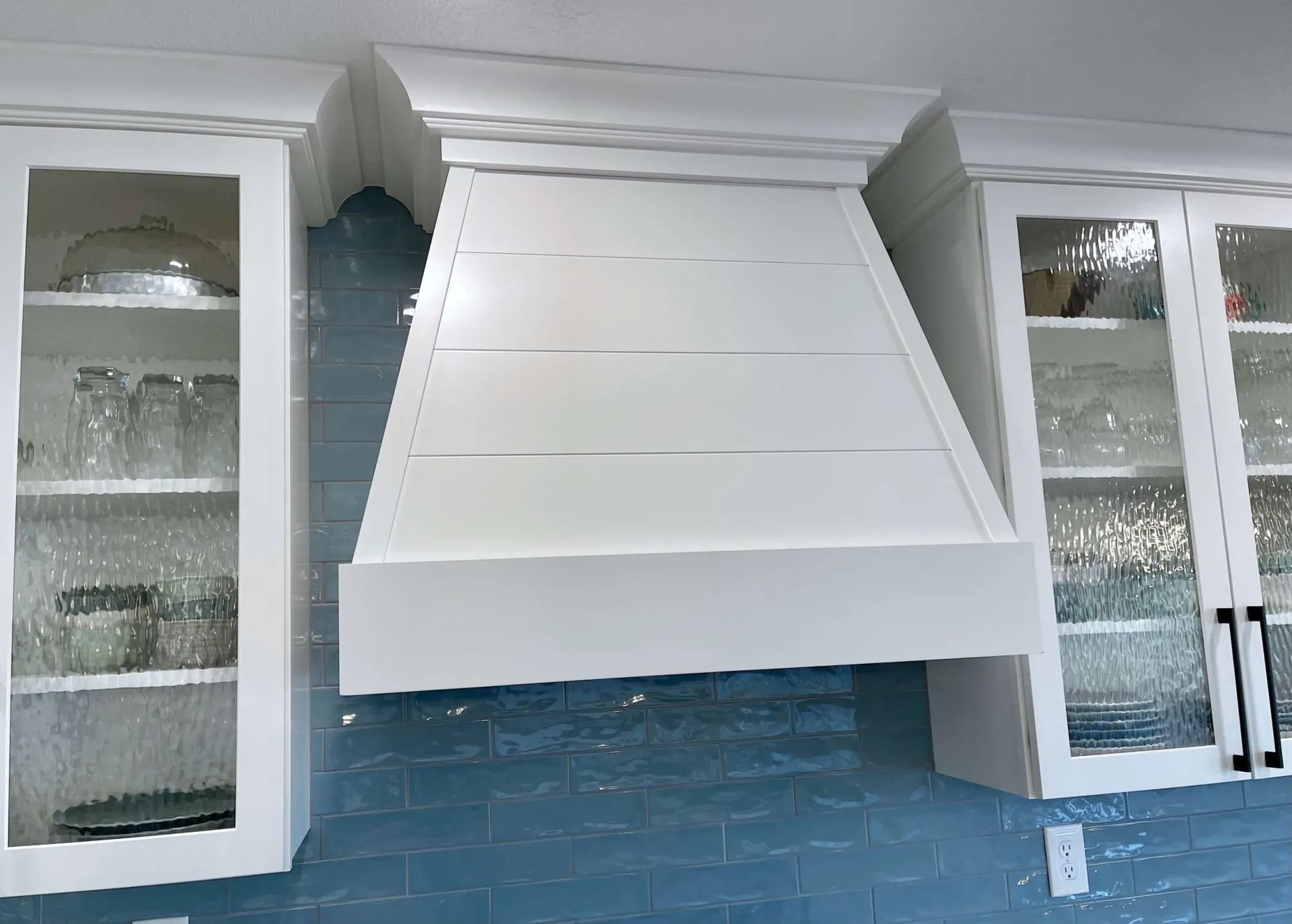 White kitchen range hood above blue tiled backsplash, flanked by white glass-front cabinets.