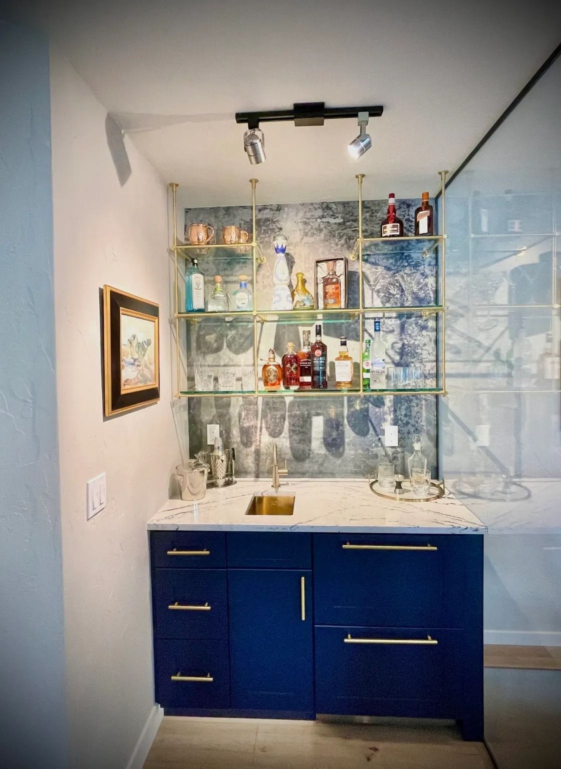 Dark blue home bar with gold shelving, marble countertop, and bottles, glasses, and a framed picture.