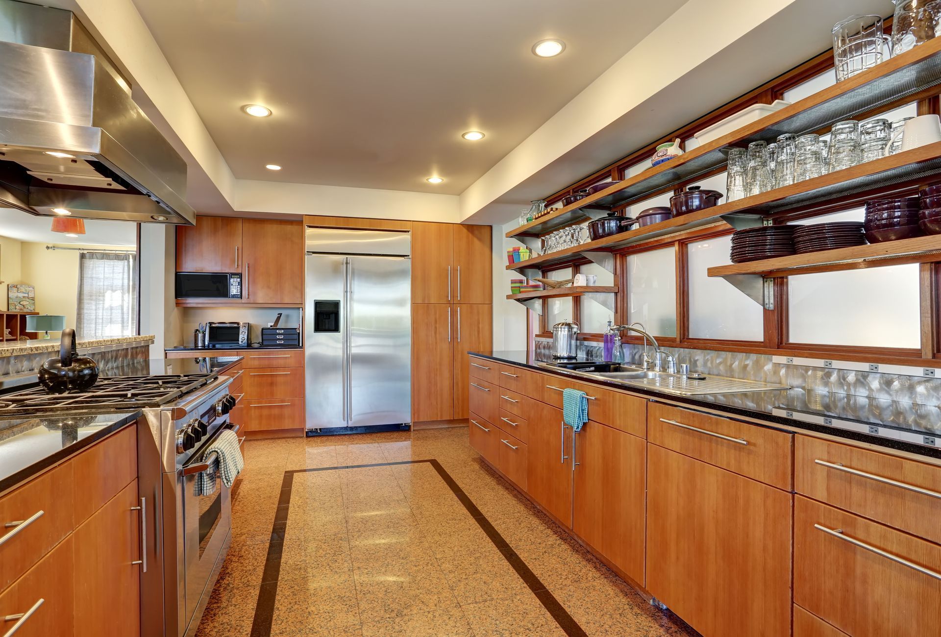 A modern kitchen features light wood cabinets, stainless steel appliances, and open shelving filled with glassware.