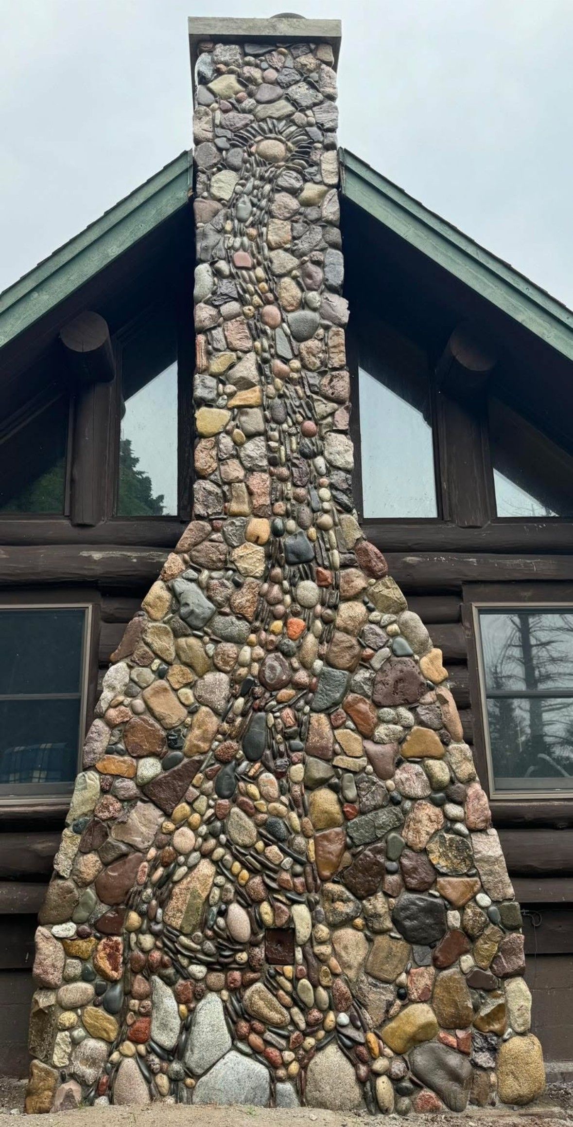 Stone chimney on a log cabin with triangular windows, set against a cloudy sky.